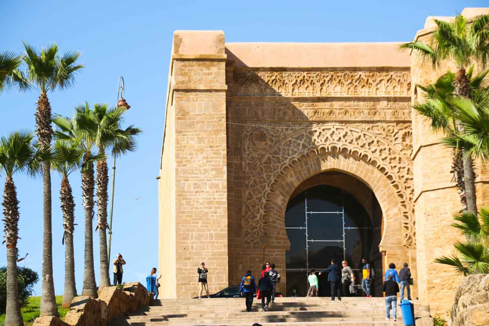 An arched stone gateway with decorative elements around and above the large arch. People in front of the gateway show that it is perhaps 5-6 storeys high. Left of it, a row of palm trees.