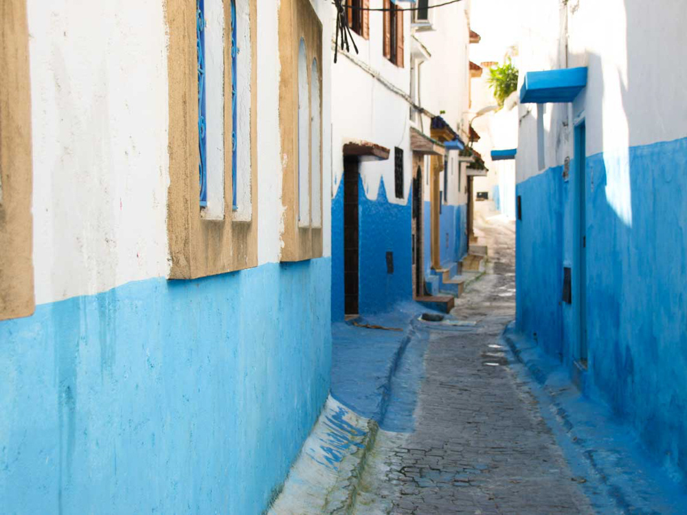 A very narrow street, only wide enough for 2 people, with whitewashed houses on either side. The houses are painted blue up to about halfway up, and white above that.