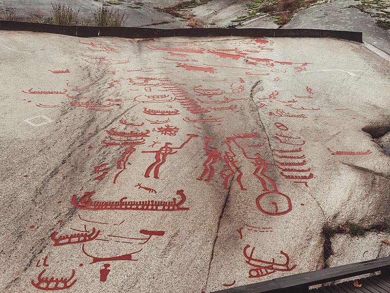 A flat, grayish-brown rock, with red line drawings on it. They portray simple boats, people, some holding sticks or tools of some sort.