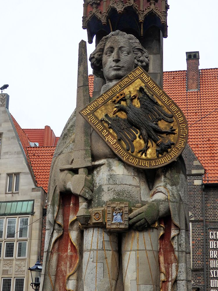 Statue of a man wearing a cloak, holding an upright sword in his right hand and with his left hand at his belt. The belt has a large center buckle with a blue-robed musician pictured on it. On his chest is a shield with a two headed eagle, a crown above the heads, and the words around the edges of the shield. 