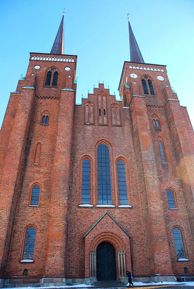Front view of Roskilde Cathedral. Red brick with two square elements that extend outward on either side of the center entrance and that support the spires above. The entrance is a rather simple romanesque arch. The large window above the entrance has 3 long vertical parts, each with a slightly gothic arch at the top. 