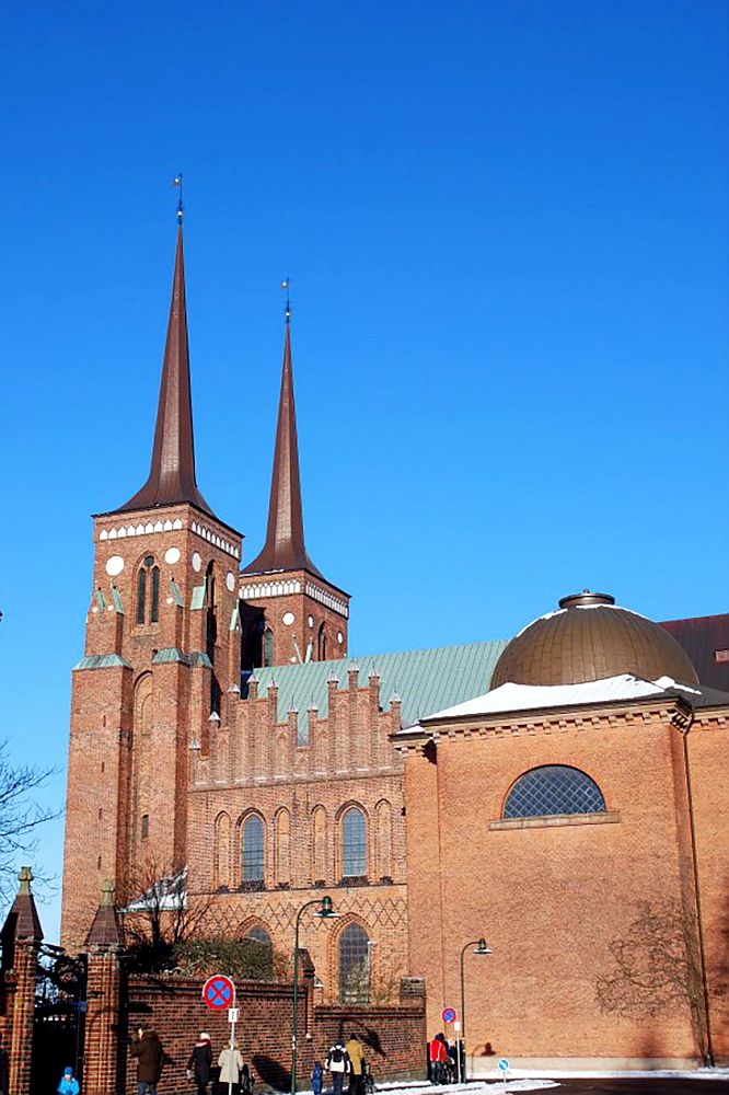 A side view of Roskilde Cathedral: 2 spires at the left, wide at the bottom but narrowing quite quickly to be very narrow for most of their length. The building is red brick with step gables on the side. A small dome is visible on the right.