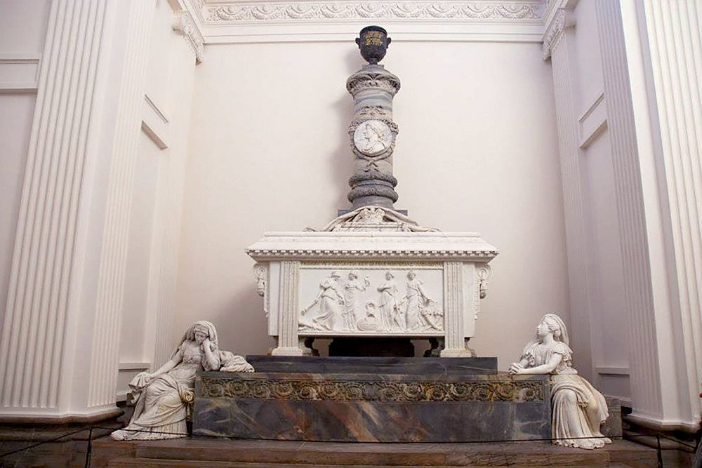 A tomb stands on a pedestal in a niche, all of it painted white as well as the wall behind it. On top of the tomb is a pillar with what looks like an urn on the top and a relief of the person's face halfway up the side. Below the tomb, a figure leans on each side of the pedestal: both are women in white marble. The left hand one leans her elbow on the pedestal and her face on her hand. On the right the woman has her hands clasped on the pedestal and looks up at the tomb.