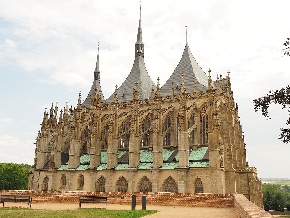 A gothic church, with a row of flying buttresses along the side, with tall gothic-arched windows between them. Three spires, not very tall and tapering to points.