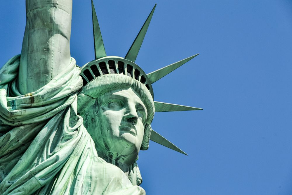 Taken from below: the face of the Statue of Liberty, with her crown with points and her arm raised.