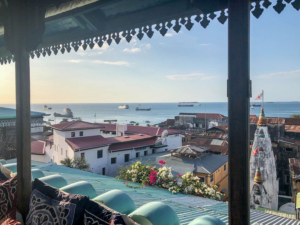 A view from a roofed porch looks out over a jumble of low buildings and one tower - perhaps a church. Beyond the buildings is the blue sea, with some ships here and there.