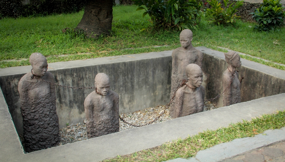 A memorial shows a rectangular pit, walled with concrete, perhaps twice the width and length of a grave, about 4-5 feet deep. In the pit are statues of five people, each up to their knees in in gravel, with hands at sides and chained together with chains around their necks. They have finely-sculptured faces, but their bodies, moving downwards, become rougher and less finished, as if they are becoming one with the gravel below.