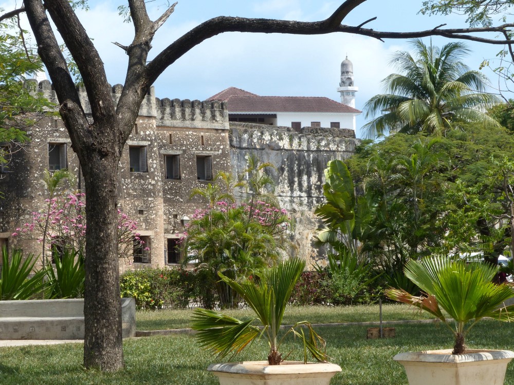 Seen as the background to a park with palm trees, the fort is made of stone walls, with crenellations along the top. It appears to be only about two large storeys tall. A white minaret peeks up behind it.