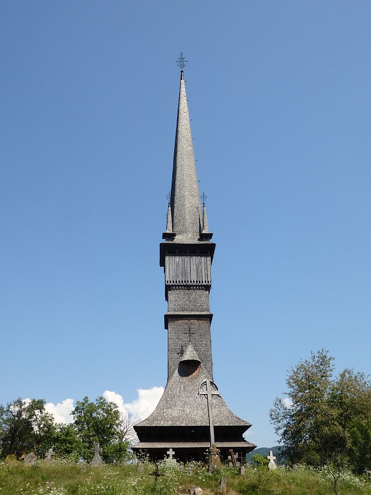 This is a particularly tall one of the wooden churches of Maramures, seen from straight on. The church's roof is very steep and shingled. From its peak, the steeple about 2 times as high as the church, so 3 times the church's height in total. The steeple is square for the lower half of its length., then round from there, and tapering to a point at the top. At the base of the round part are 4 little turrets toped with crosses.