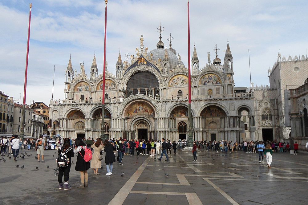 St Mark's Basilica as seen across a flat plaza with tourists. Big arched gateways into the building and a lot of small towers or turrets around one big central dome.