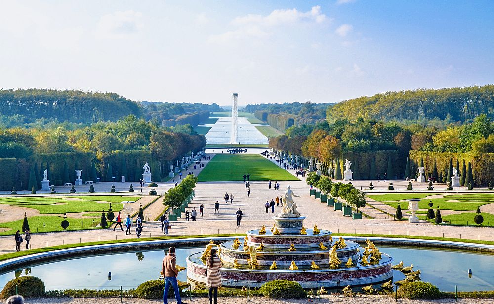 In the foreground, an ornate round fountain like a multi-tiered wedding cake, each ring with gold statues and a marble statue on the top. The fountain sits in a round pool. Beyond that, a long view down a long "road" with forests on either side. The road is grass with statues lining the sides. In the distance, continuing the road is a long rectangular pond, with a tall statue of some sort in it. 