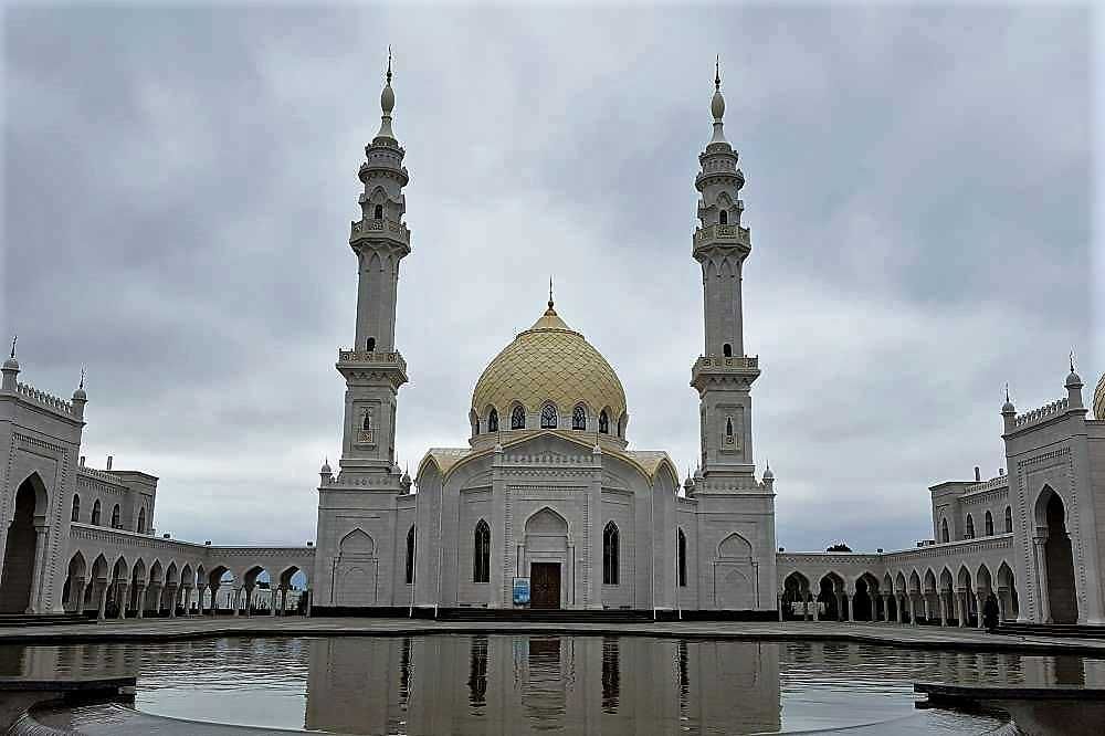 In the foreground, a pond. Beyond the pond, a mosque: very symmetrical, with a gold dome on its top and two tall minarets, one on each front corner of the building. Porticos extending out both sides of the building.