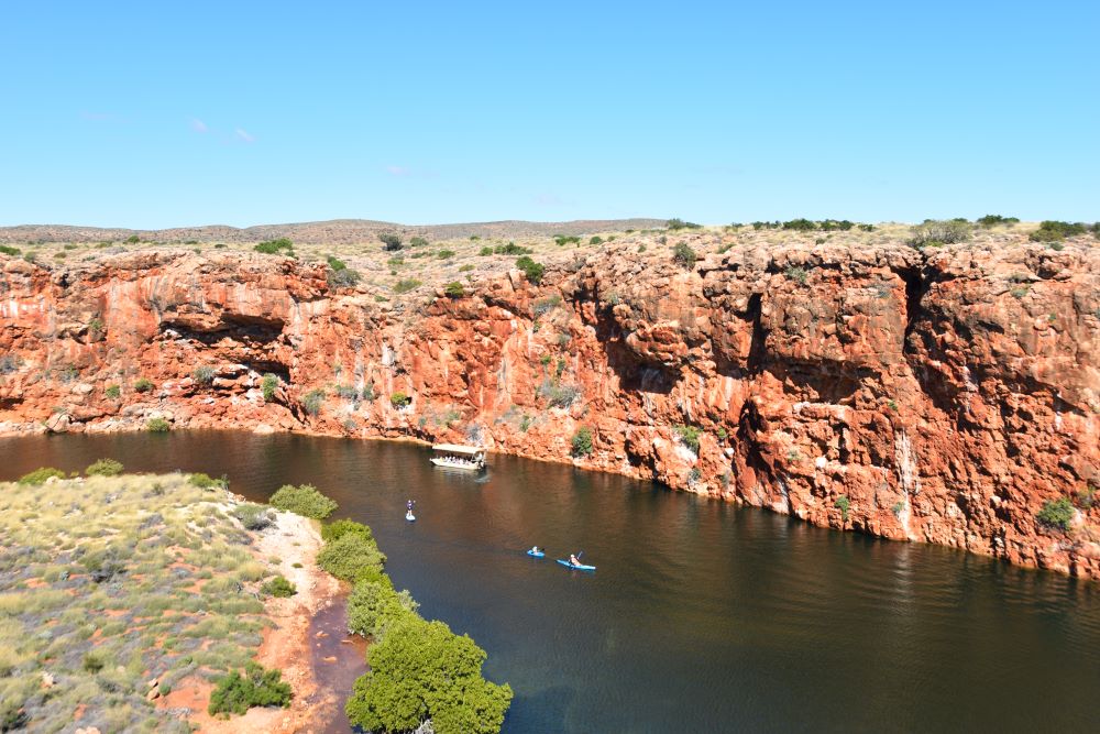 A view into a gorge with, on the near side, a low grassy bank, but on the far side, a very high vertical wall of red stone. A small boat and several kayaks are visible on the river that runs through the gorge.