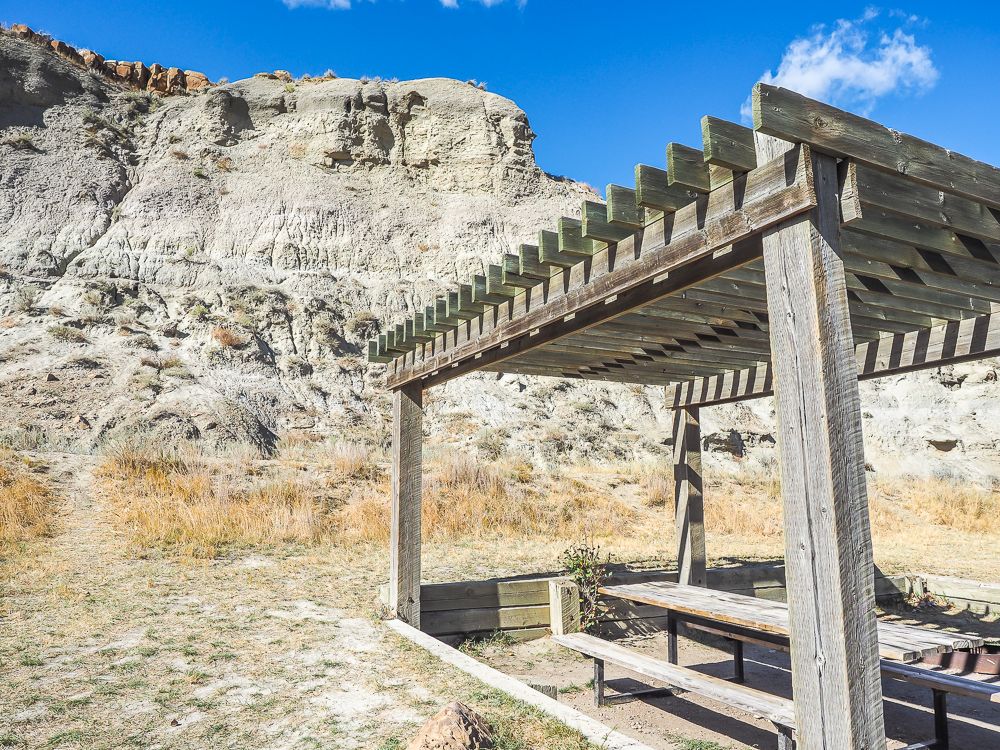 The side of an eroded hoodoo in the background. In front of it, a wooden shelter with a picnic table under it.