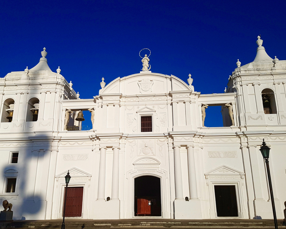 Front view of Leon Cathedral: white plaster, symmetrical and only two storeys, wider than it is high. An arched roof in the center, squat turrets on either side. A statue center top of the building, classical decorative elements above each door and window.