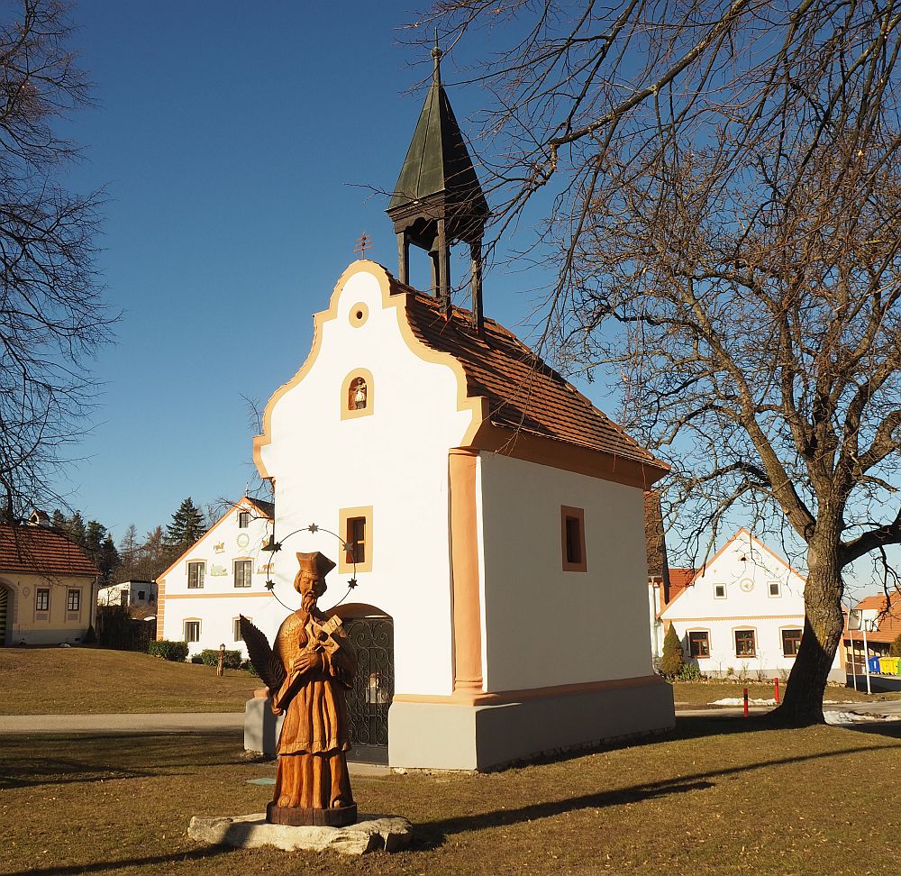 The chapel is very small and simple, with a single small window in each wall and a small spire. The front has a decorative outline to the gable. In front of it is a statue of a saint in robes, holding a cross.