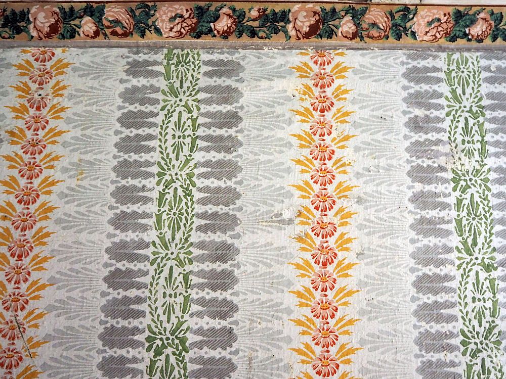 Vertical repeated patterns, each vertical a different color: from left to right: orange and red flowers, light gray leaves, darker gray leaves, green flowers and leaves, back to the dark gray leaves but printed in the other direction, back to the light gray, but again reversed, then back to the red and orange., and so on. Above a piece of the molding can be sean, hand-painted with pink roses and green leaves between them.