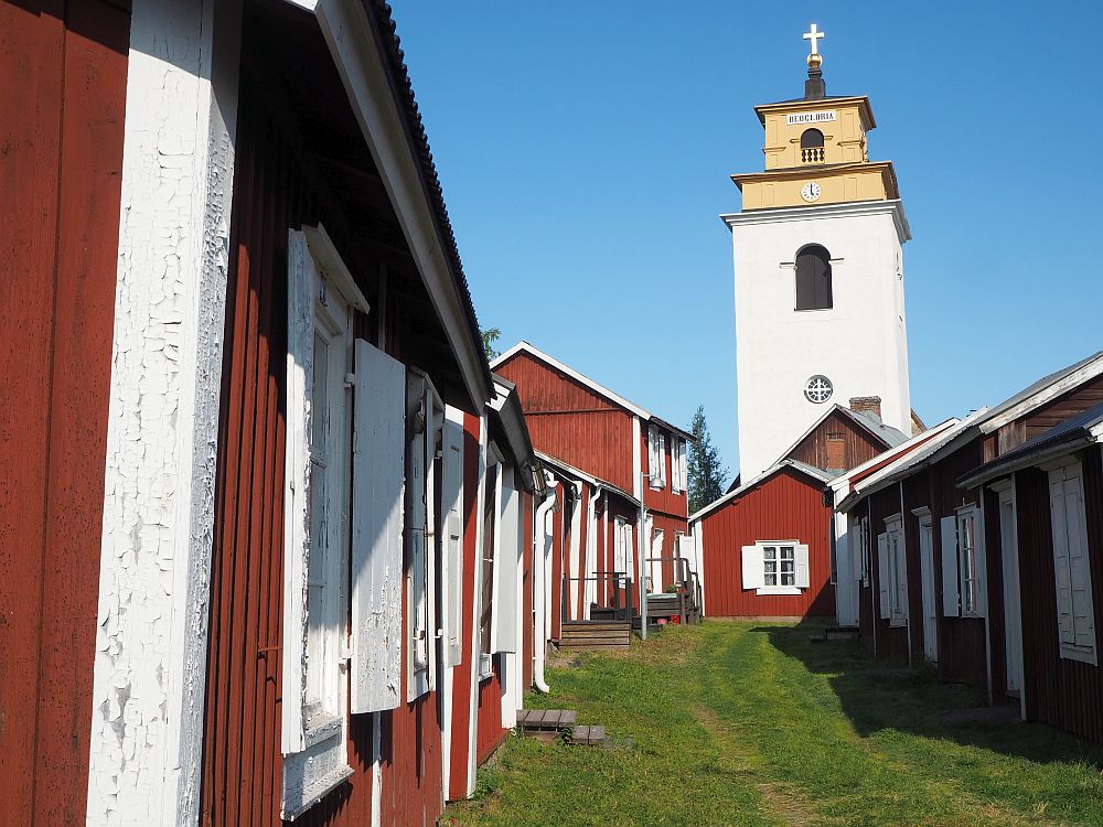 looking along a "road" between two rows of small red houses with white trim. The road is grassy. At the end of the road is a tall white church tower.