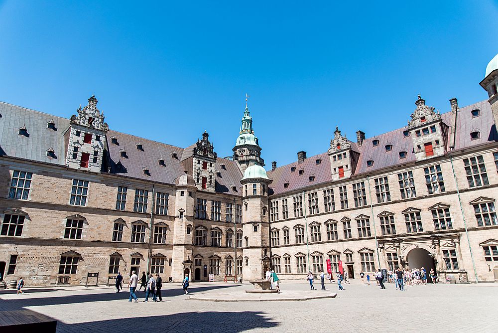 two sides of the inner courtyard of Kronborg castle. Walls are mostly flat with many windows on each story: 3 stories under the roofline, perhaps 2-3 more under the roof, which has large 2-story dormers. In the corner is a small tower with a green copper roof. People enter through an archway in the right-hand wall.