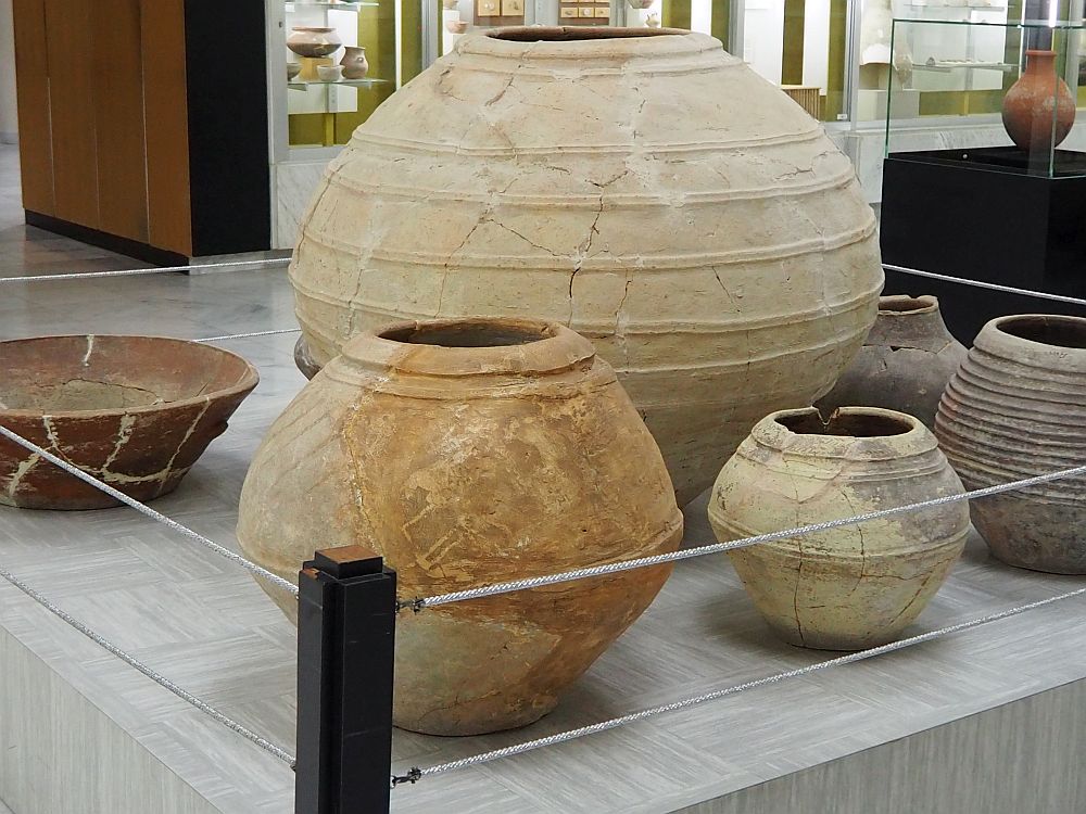 One very large storage pot of clay, surrounded by five more of various shapes and sizes, but all less than half the height of the tall one. They are all very simply-formed, with just lines or ridges around them for decoration.