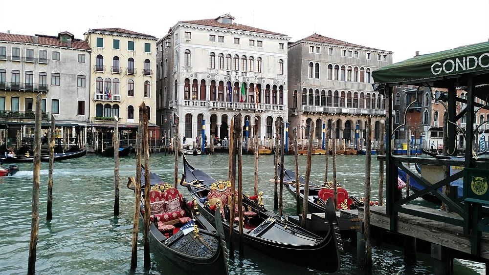 Foreground: gondolas "parked" between poles sticking up out of the water. The water is greenish and on the other side of the canal is a row of elegant buildings, mostly with arched windows on the water-level floor and the couple of floors above that. Each building has a relatlvely flat top and 4 stories in total.