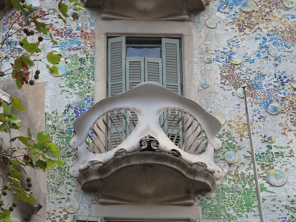 A small balcony in front of a window. The window is shuttered with light green shutters. The balcony has rounded forms and wavy edges like an organic form. The wall around the window seems to be scattered with mosaic tiles - mostly quite small, but some are larger and round - in blues and greens.