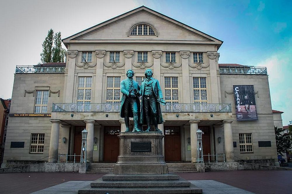 A grand building, almost symmetrical, with a statue of Goethe and Schiller in front of it.