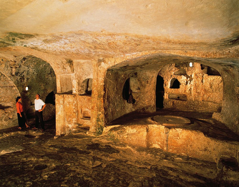 A low-ceilinged room, carved out of rock. At the back are two archways (carved, not built). Two people stand in the left-hand archway, showing that the height is about twice a human's height. The right-hand one is larger, and through it more rough-hewn space can be seen in another "room."
