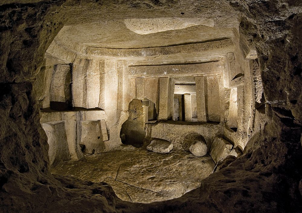 A view framed by a stone wall opening looking into a room carved in stone. It looks like the ceiling has been carved to look like it has beams holding it up. Across the room is a doorway of sorts, but in the middle of the wall, not to the floor, and it shows a small part of a room beyond this one.