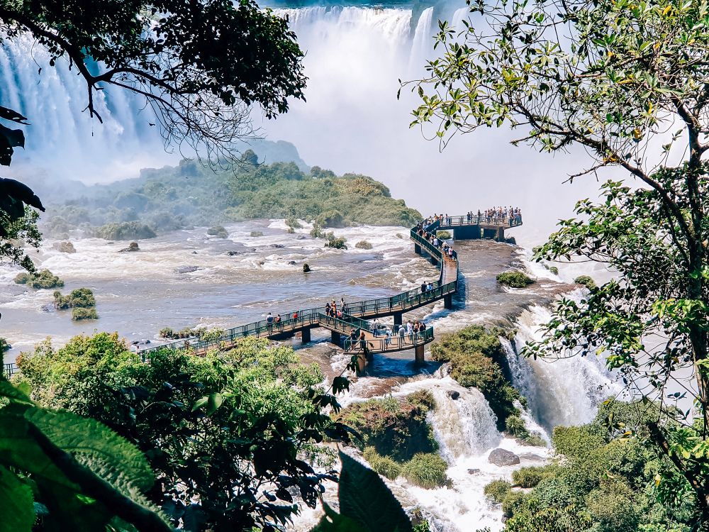 A view framed by trees overlooks the wooden walkway with rails that passes over a relatively flat part of the Iguaçu falls in Iguaçu National Park. The walkway ends at a viewpoint right on the edge of the waterfall, but the length of the walkway also edges part of the falls.