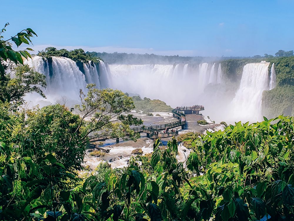 A view of part of the waterfall: the tops of trees in the foreground, in the middle distance a wooden walkway winds over a flat part of the falls, in the background a wall of white falling water coming off a wide flat cliff.