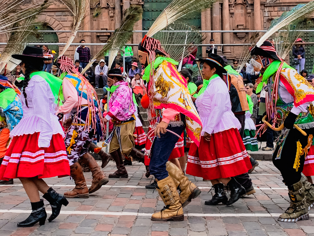 Traditional dancers, all facing to the left. The women have wide red skirts and white blouses and wear short boots and hats with wide brims. The men wear black pants tucked into high boots. They wear a long shawl down their backs, tied in front and a hat with tall feathers sticking out.