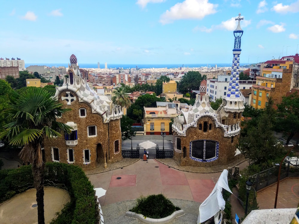 In this partial view of Park Guell, two buildings are visible against a much wider view of the city and the sea. The two buildings are brown stone with colorful roofs that make them look like something out of a fairy tale: then come to a point in a series of wavy shapes. The house on the right also has a tall spire in blue and white tiles with a cross on the top.