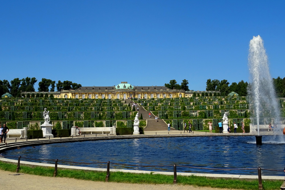 View of the Sanssouci Palace in the background on a low hill, with, in the foreground, a large pond with a fountain in the middle of it.