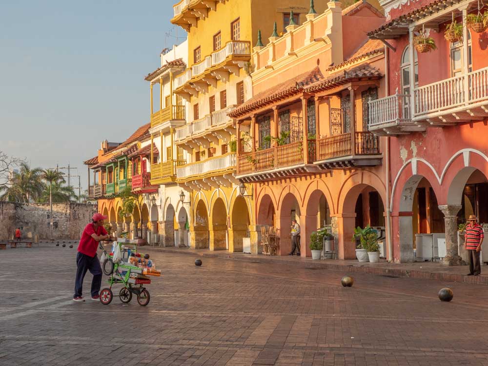 A plaza in the foreground. Right and behind, a row of colonial-era buildings painted in pastel colors. Each is from two to four stories tall and all have balconies above the ground floor. The ground floors have arched colonnades shading the sidewalk. On the plaza is a lone vendor pushing a small wagon.