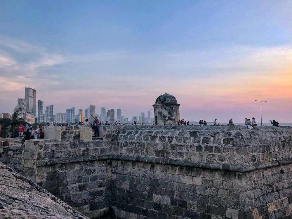 In the foreground, stone walls with people walking along the tops. In the background the modern city skyline with its skyscrapers.