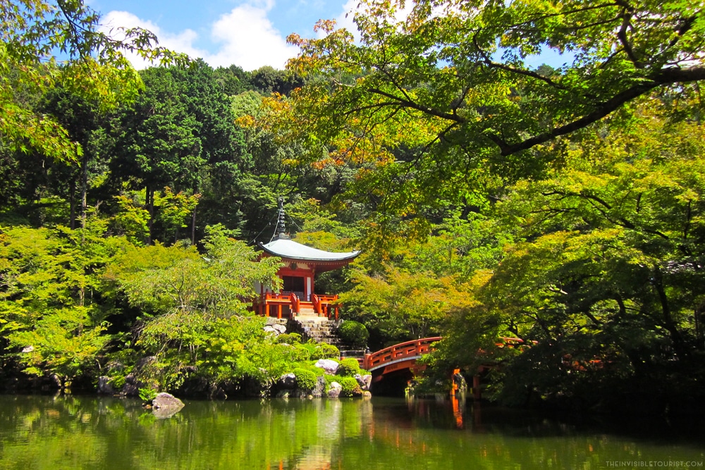 A small red-painted temple and arched red-painted bridge peek out from amidst the green foliage on the edge of a body of water.