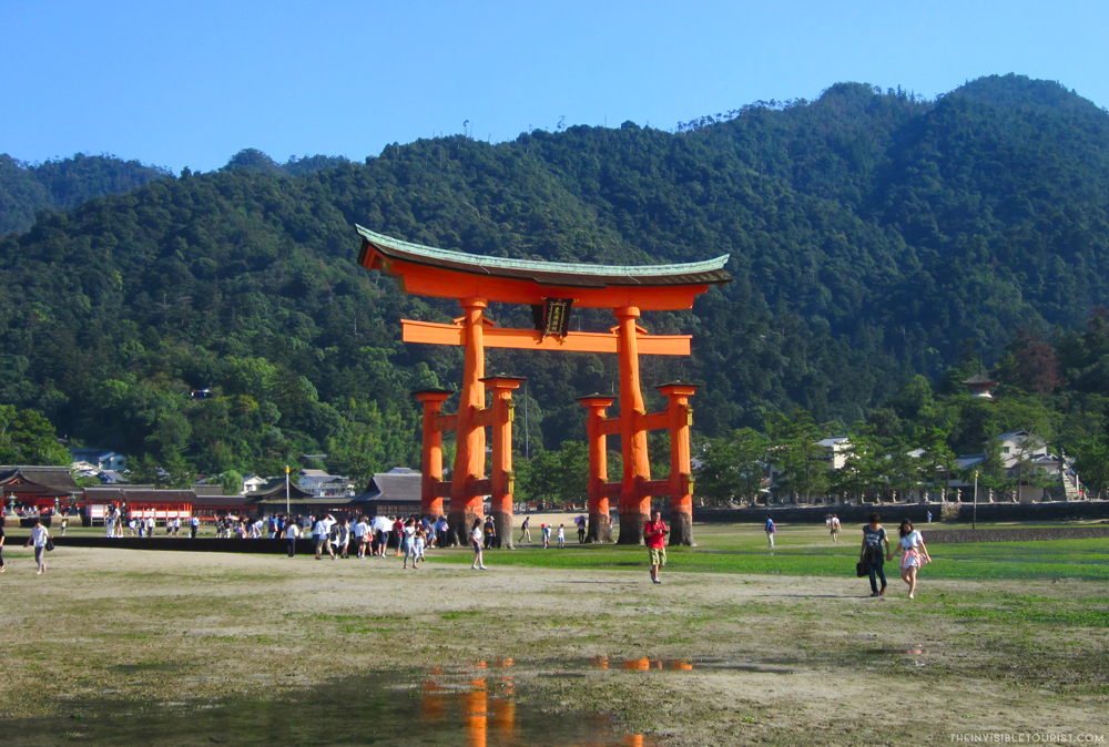 The Torii gate stands on two massive poles, each with two smaller poles flanking it. A lower, straight crosspiece near the top, and along the very top, a crosspiece that is slightly curved upward at each end. The whole thing is painted reddish-orange. It stands in a flat field, with a wooded hill behind.