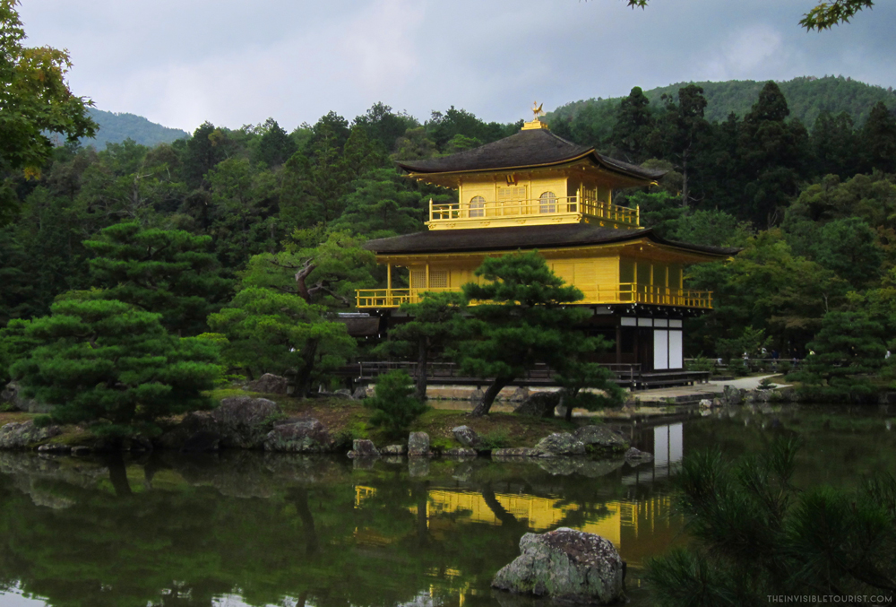 A bright yellow temple with curving brown roofs on each story, standing on the shore of a lake.
