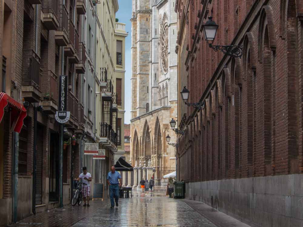 Looking down a street between brick buildings, the cathedral is visible at the end of the street, its facade at an angle. What is visible is three archways on the ground floor, and it looks like a round gothic window above that.