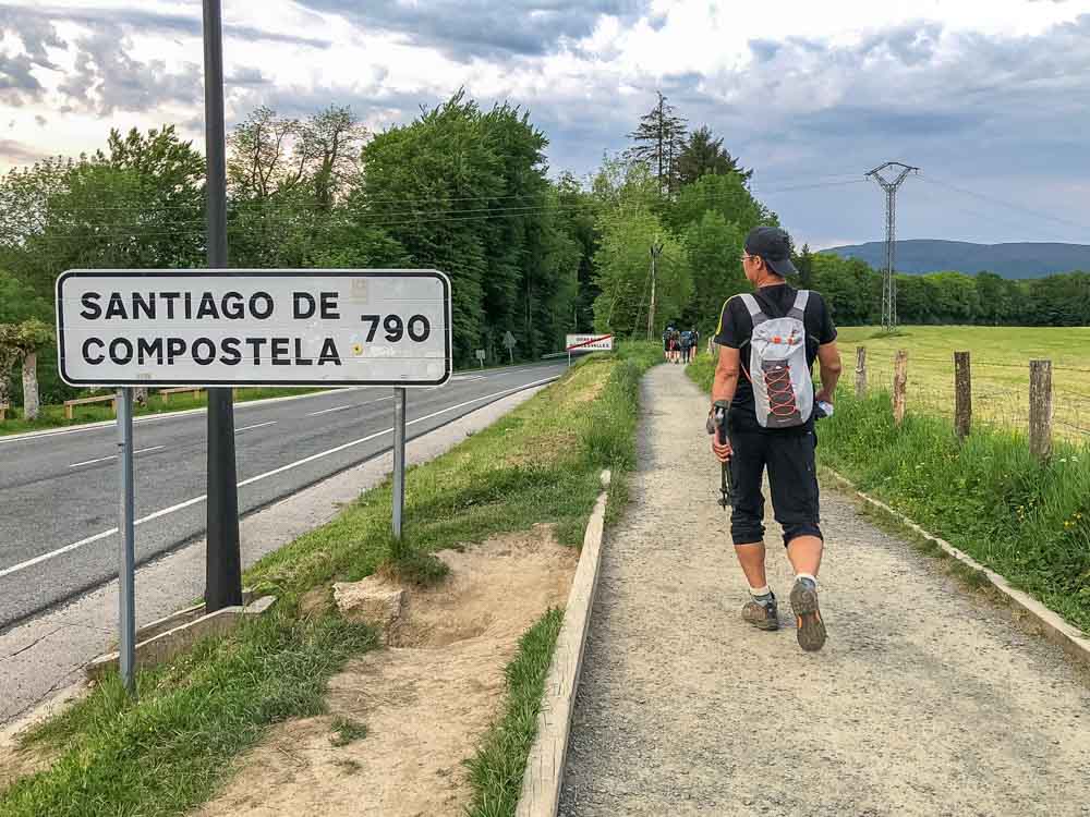 A young man dressed in walking clothes and carrying a small backpack passes a sign reading "Santiago de Compostela 790"
