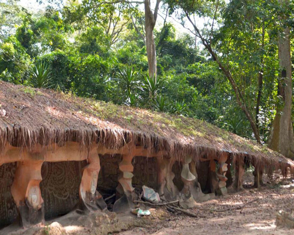 A long low building with woods behind it. The building has a thatched roof that is held up along the front by a row of stubby pillars, each of which is carved abstractly.
