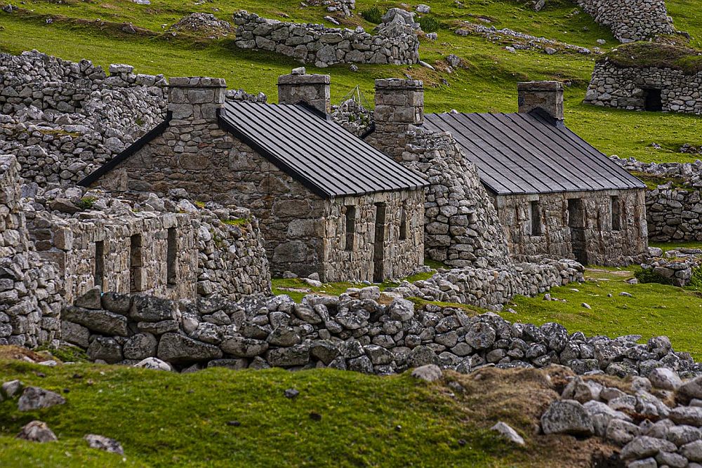 Two of the restored houses on the "main street" on St. Kilda. They're very simple stone-built single storey houses, with a peaked roof and a chimney at either end and a door in the middle of the front, one window on either side of the door. Lots of ruined low walls around the houses and one unrestored house next in the row: similar front wall but no roof.