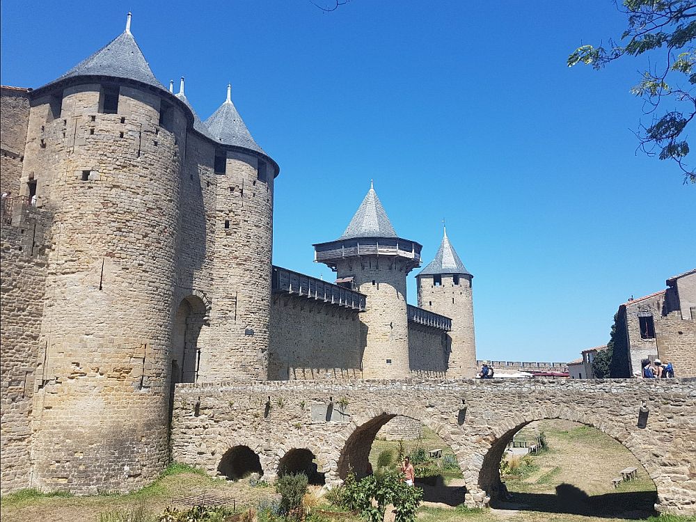 The fortress walls on the left, with rounded towers flanking a gate. A stone bridge with multiple arches leads into the gate. Along the wall are two further round guard towers with pointed roofs.