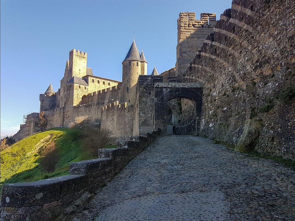 A road along the edge of the fortifications: wall on the right, drop-off to the left. Straight ahead: walls with crenellations, a round tower, and part of a stone castle with a square tower.