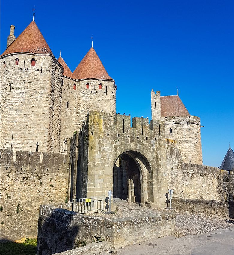 An entrance through the fortification walls has a large arched gateway. Beyond that is a castle, with high, mostly windowless walls, made of brick or small stones. The pointed roofs are reddish.