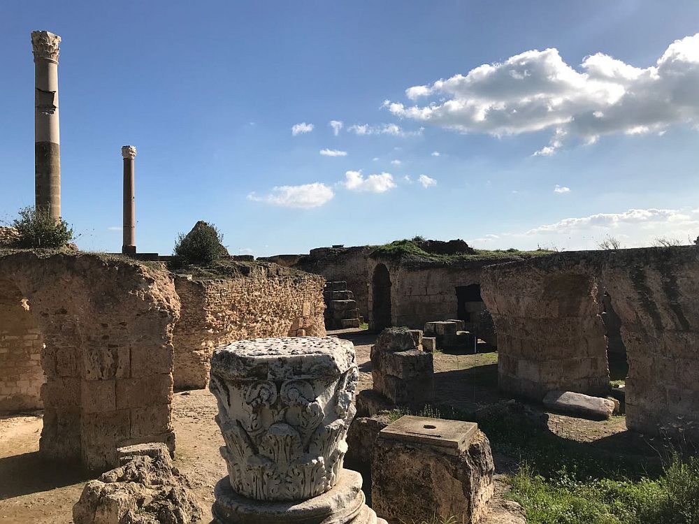 Among the ruins: various fragments of stone walls and pillars, with two tall pillars in the background. In the foreground is one ornately-carved Corinthian pediment.