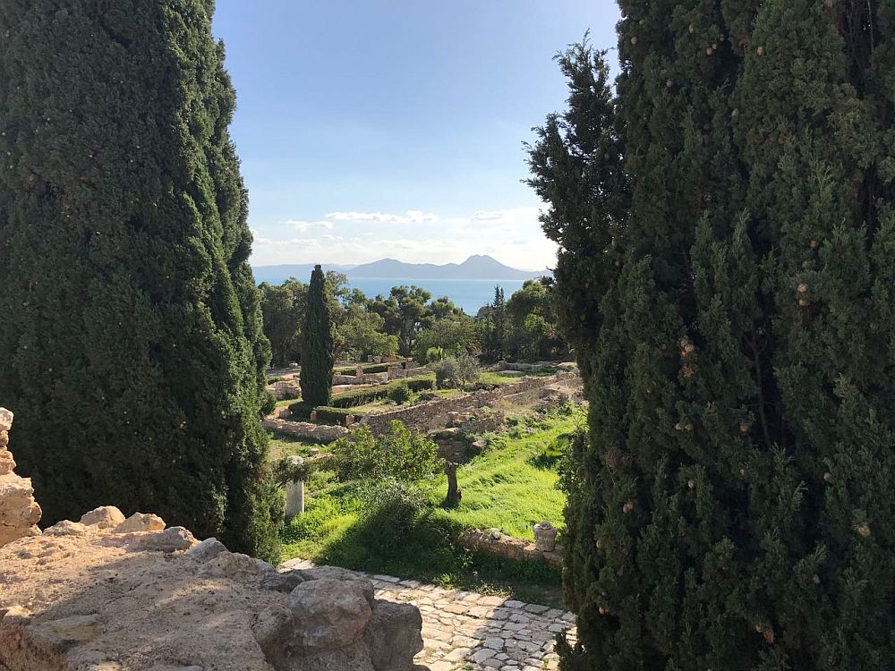 Looking from some height between two cypress trees, onto a field with ruins of a building. Low walls of stone are all that is left. In the distance, blue water and mountains beyond it.