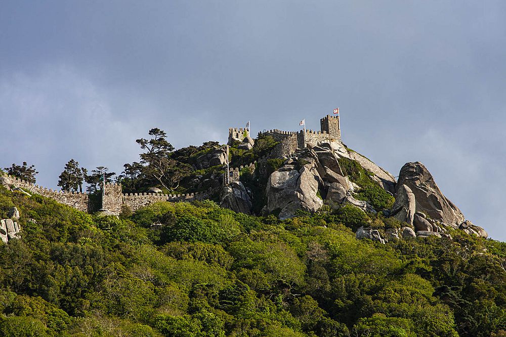 Looking up at a rocky hill, the walls of a fortress or castle are discernible along the heights, with crenellations and watchtowers.