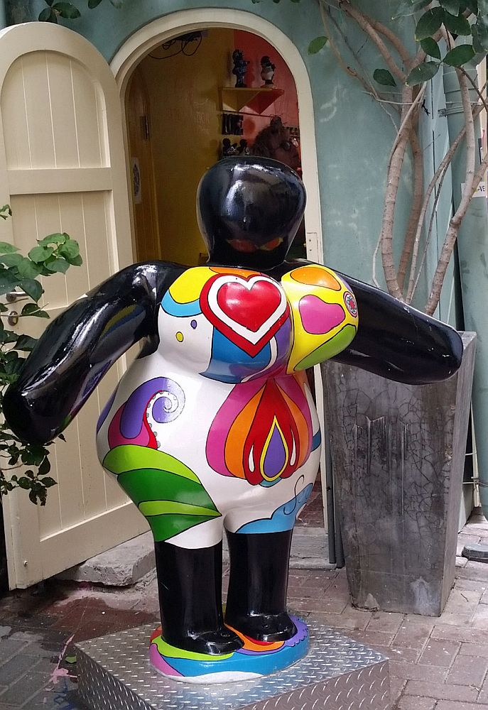 A larger-than-life "Chi-chi" doll standing in front of a doorway. It is shaped like a very rounded, very buxom woman. She is painted in multi-colors: a heard on one breast, lots of colors on the other, etc.